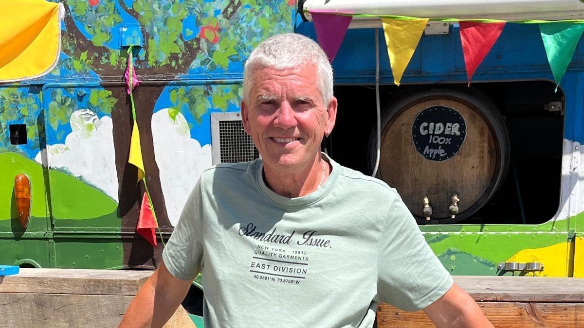 Dave smiles at the camera in front of a decorated caravan with bunting across the side. A keg of cider can be seen inside the caravan. He has grey hair and wears a light blue t-shirt.
