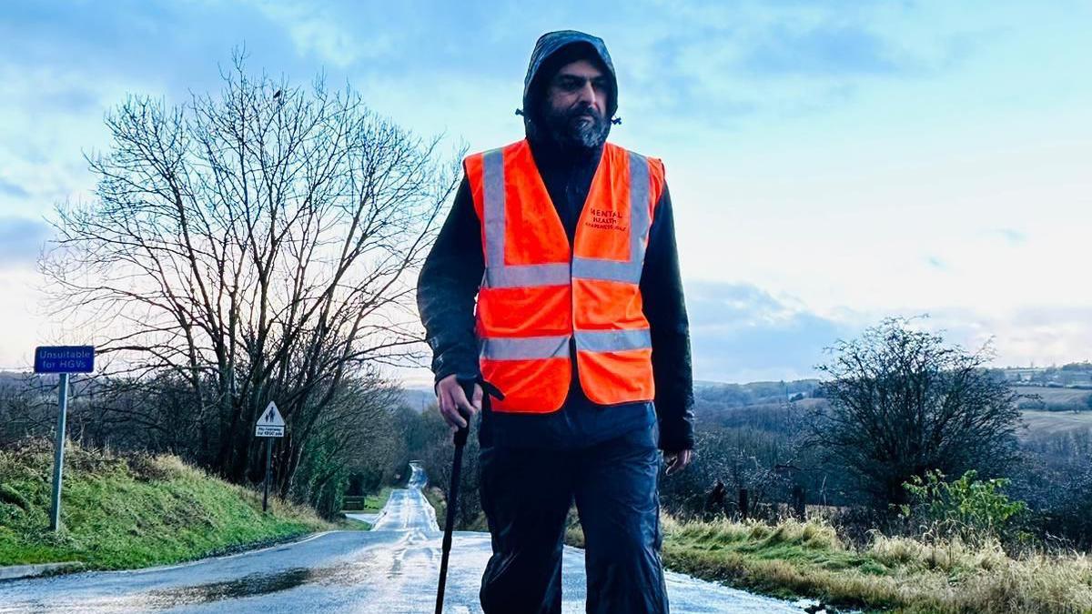 Bhupinder Sandhu is wearing dark waterproof clothing with his hood up, along with an orange high-visibility waistcoat. He is holding a walking pole as he walks along a quiet country road with trees and grass verges on his walk between England and Scotland. There are fields in the background.