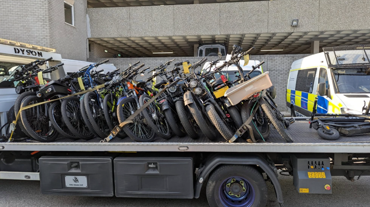 Recovery truck with e-bikes on the back of it. It is a large vehicle and parked in front of a Devon and Cornwall police van.