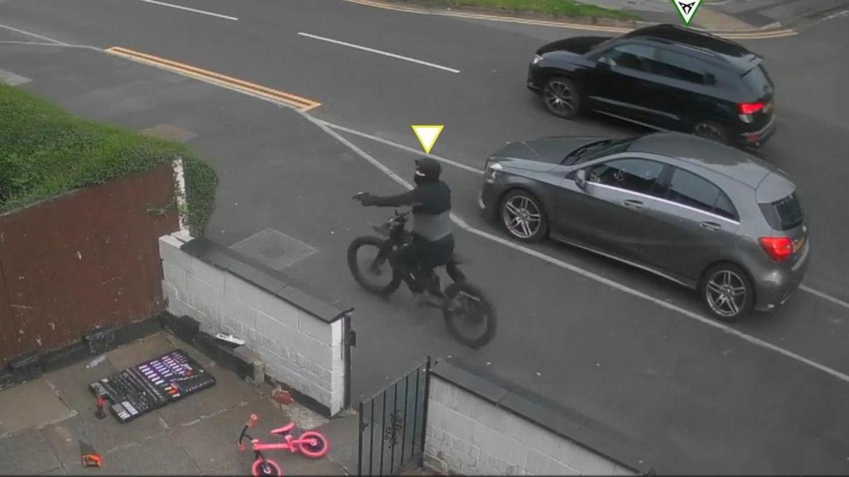 A man dressed in a black and grey coat, wearing a black face-mask, sits on an electric bike pointing a handgun towards the front-yard of a house. A child's pink bike and an open tool-box are visible in the yard area. 