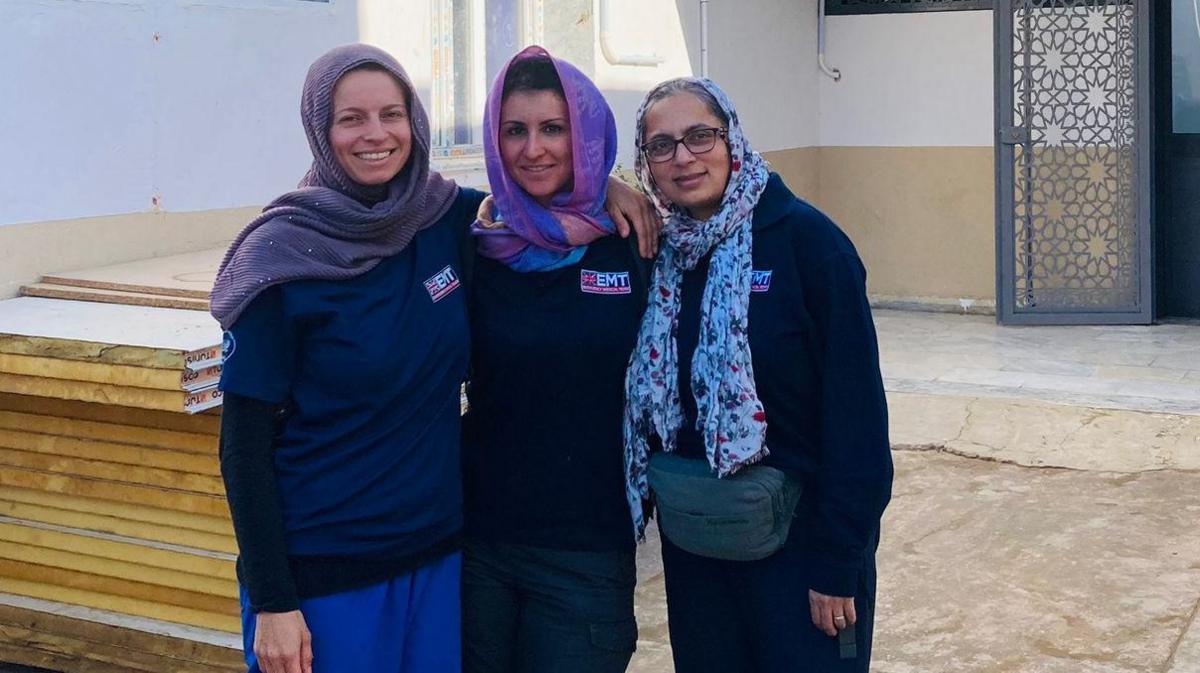 Alessandra Morelli is standing between two other women. All three women are wearing headscarfs. They are standing in front of a large pile of wood that appears to be being used in construction. There is a a cream building behind them. 