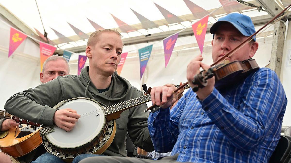 Two men. The man on the right is wearing a blue shirt and cap, and is playing a fiddle. The man on the left is wearing an olive-coloured hoodie and is playing a guitar-like instrument. 