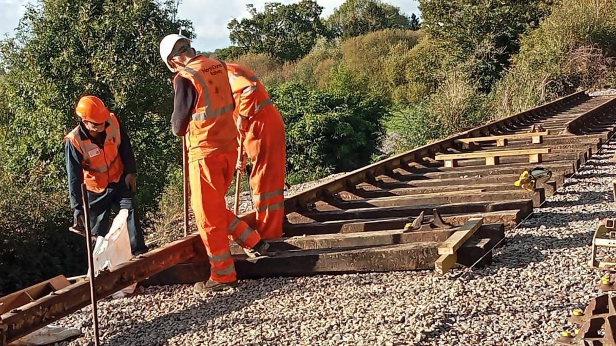 Three men in orange hi-viz overalls and hard hats manoeuvring a section of steel railway track into position. Behind them a new section of track and sleepers are already in place.