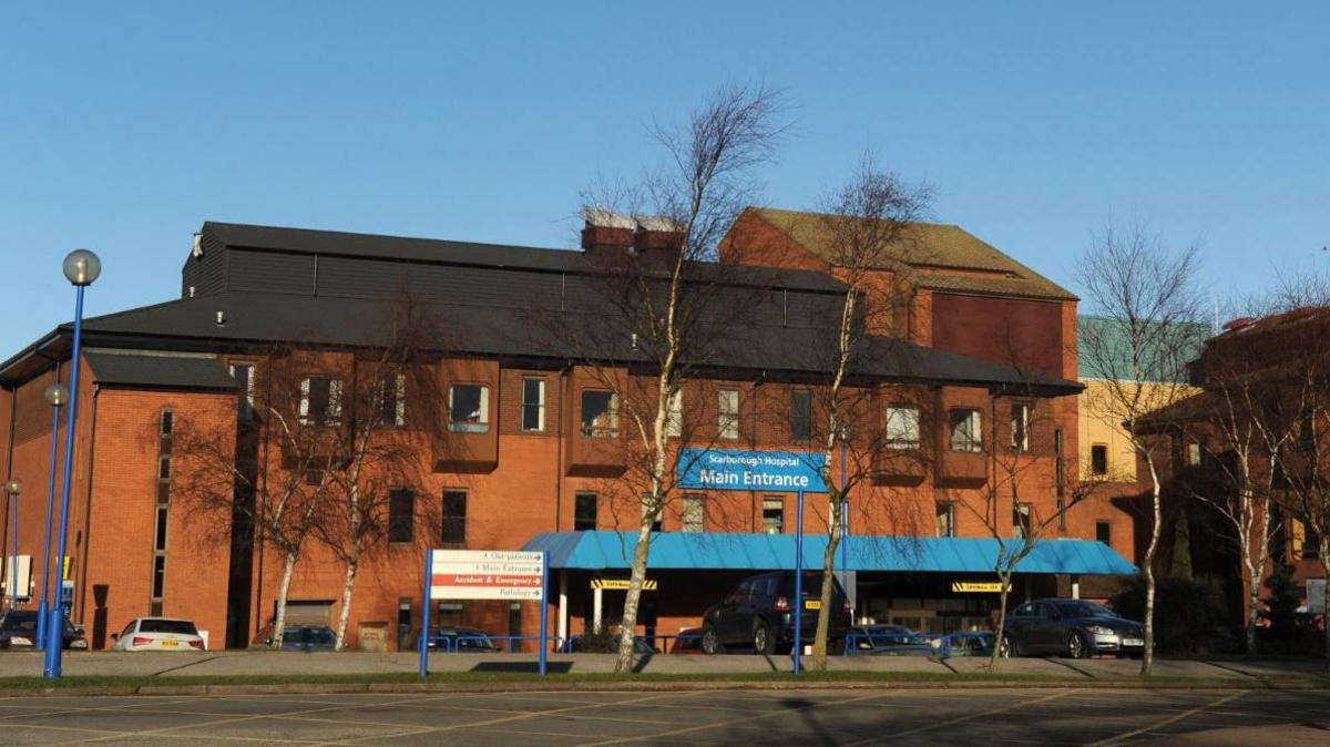 Main entrance to Scarborough Hospitals: a modern red-brick building, with a blue awning on a tree-lined street