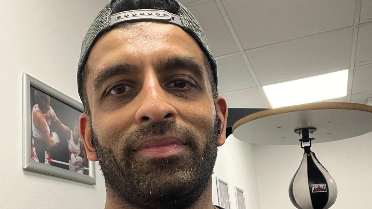 Harry Patel wears a backwards facing cap and smiles at a camera inside a gym. 