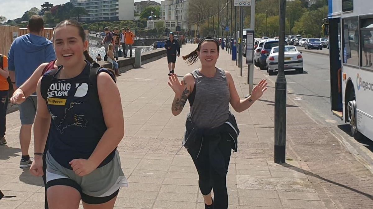 A smiling Jasmine runs along the sea front with a fellow runner smiling alongside her 