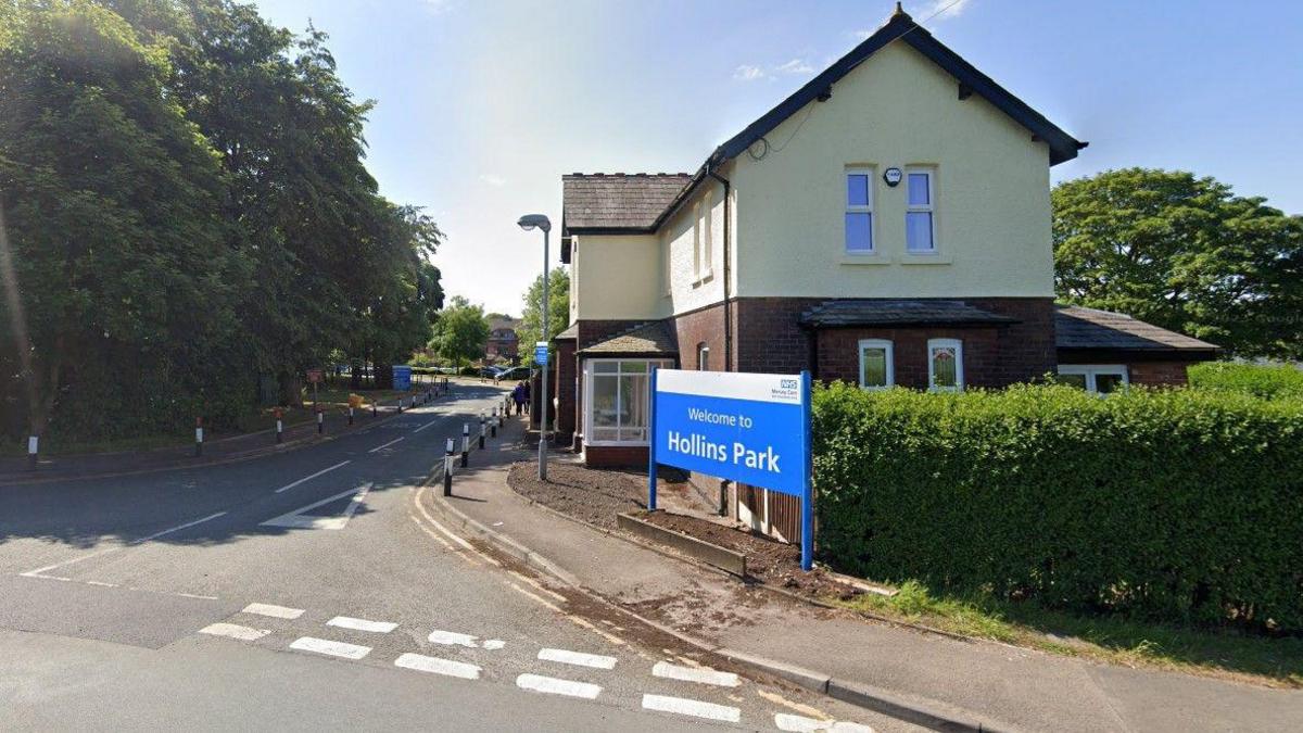 A blue and white sign at a junction, in front of a detached building, which says Welcome to Hollins Park