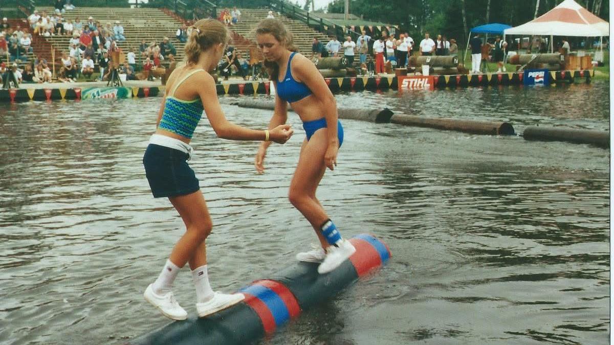 Two women with their dirty blonde hair pulled back, one in a two-piece swimsuit and the other in shorts and a tank top, balance in socks and sneakers on a rolling log in water as stands of spectators look on