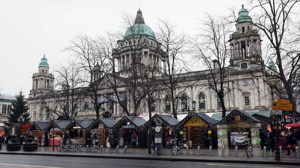 Stalls outside of Belfast City Hall at the Christmas markets. There are Christmas lights up and the weather is damp and grey. 