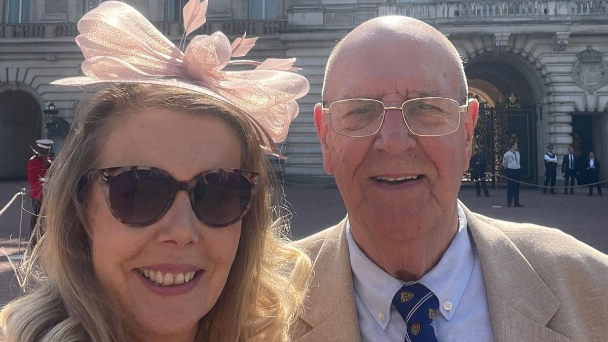 A male and female are taking a selfie - they are both smiling directly at the camera. The woman is blonde, wearing large tortoiseshell sunglasses and a pink fascinator. The male is bald, wearing thin silver glasses and a beige suit jacket with a blue shirt and navy tie.