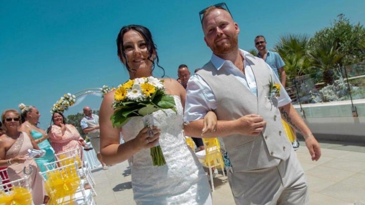 Daniel Hendron on the right with a brown beard and sunglasses resting on the top of his head. A woman and Mr Hendron are arm in arm, with the woman carrying flowers in her right hand. Other people are in the background.