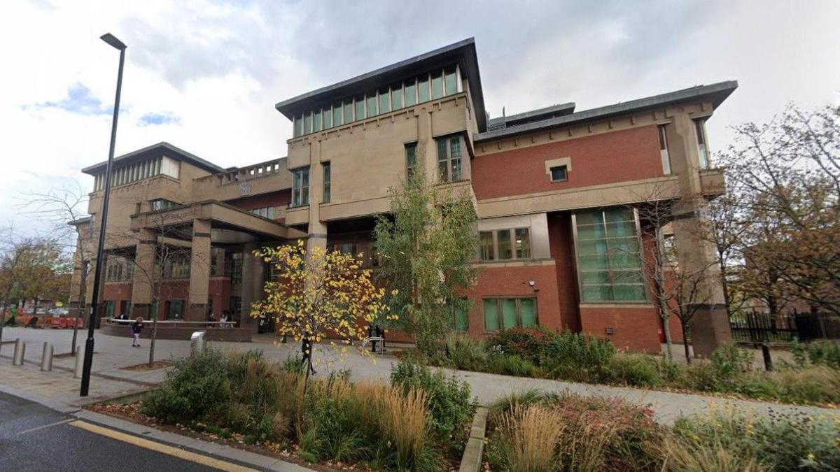 Exterior of a modern red brick and yellow stone building with square columns, differently sized windows and a flat roof. In front of it is a pavement and in front of that is greenery at ground level and a tree with yellow leaves. Above is a cloudy sky.