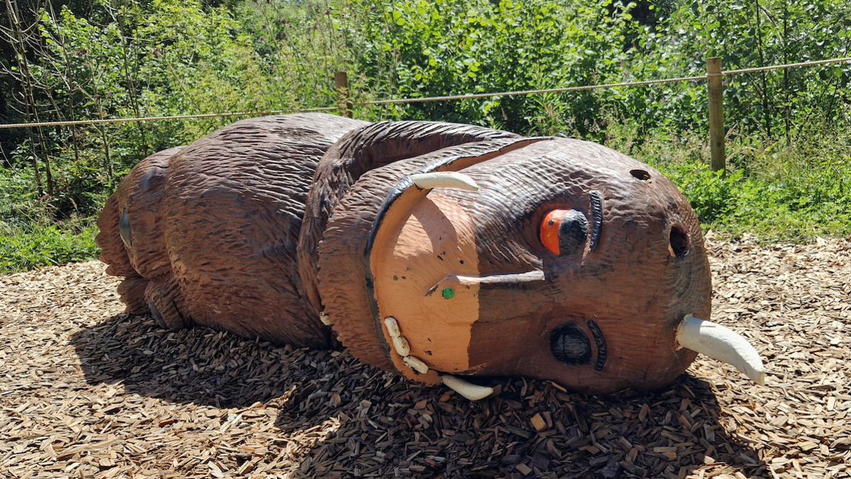 A large wooden sculpture of the children's book monster - the Gruffalo - shown lying on its side in a forest with a horn and ear missing