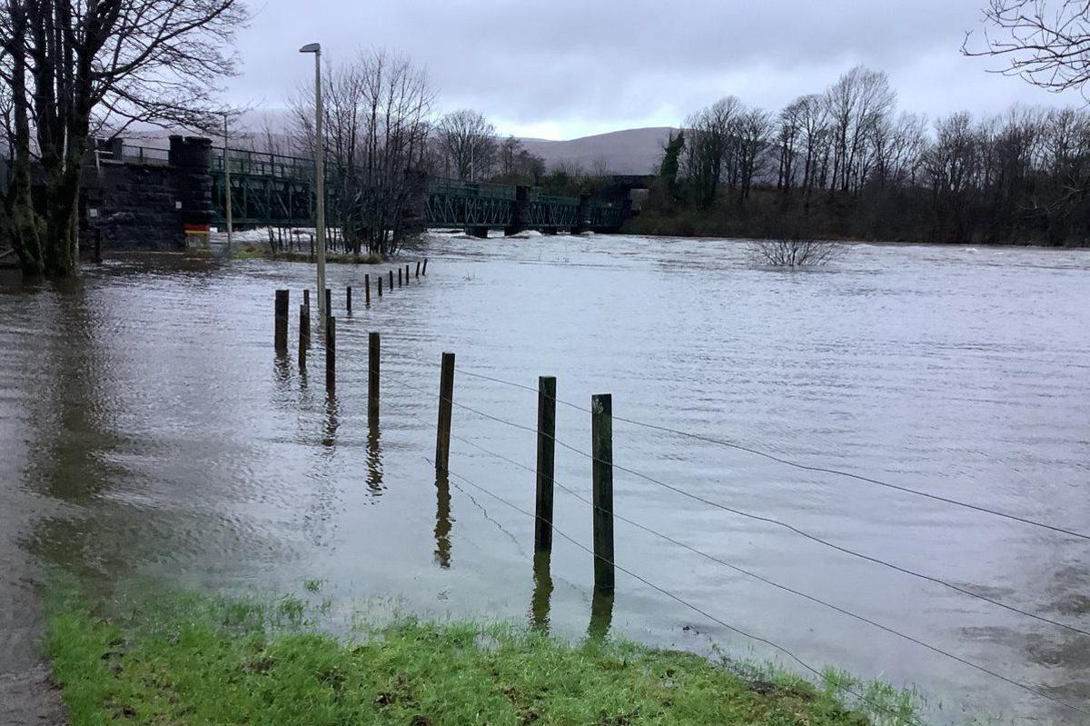 Flooded river with fence posts and a lamp post sticking out the water, trees and hills and bridge in background.