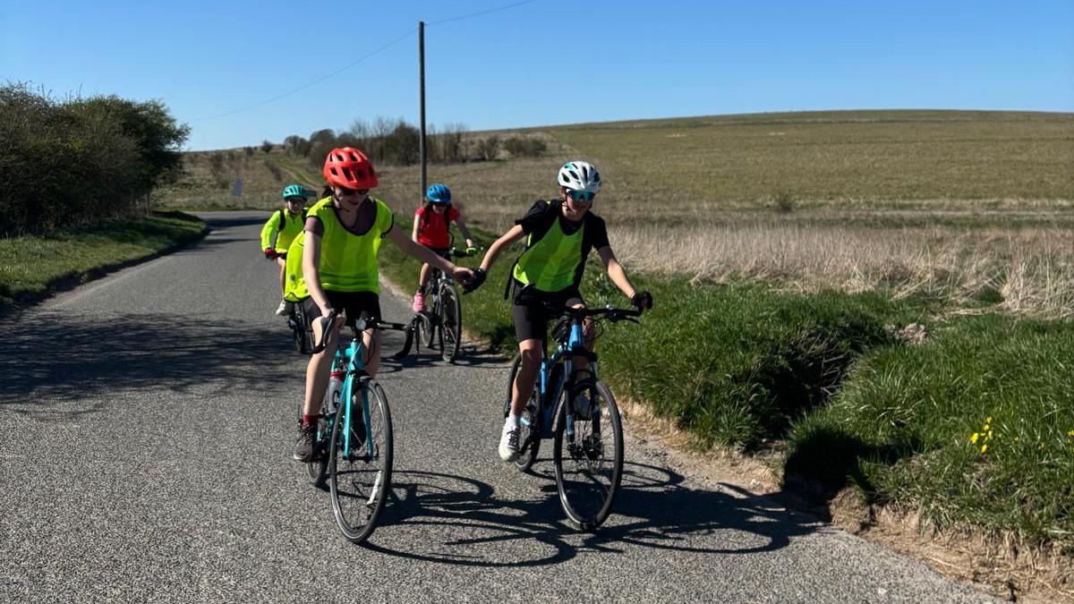 Four students riding bicycles along a road. None of them are looking at the camera, but at the bikes or ahead of them. They are all wearing helmets and biking gear.