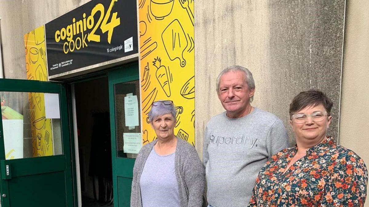 Jane Patricia Sinclair, Paul Scofield and Carol Williams standing outside a building holding Cook24 sessions. 