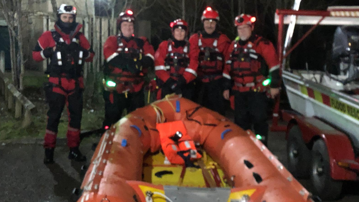 Five members of the Cornwall Surf Life Saving Clubs are wearing wet weather gear, helmets with lights on and buoyancy aids. They are smiling and standing behind an inflatable boat and it is night-time. 