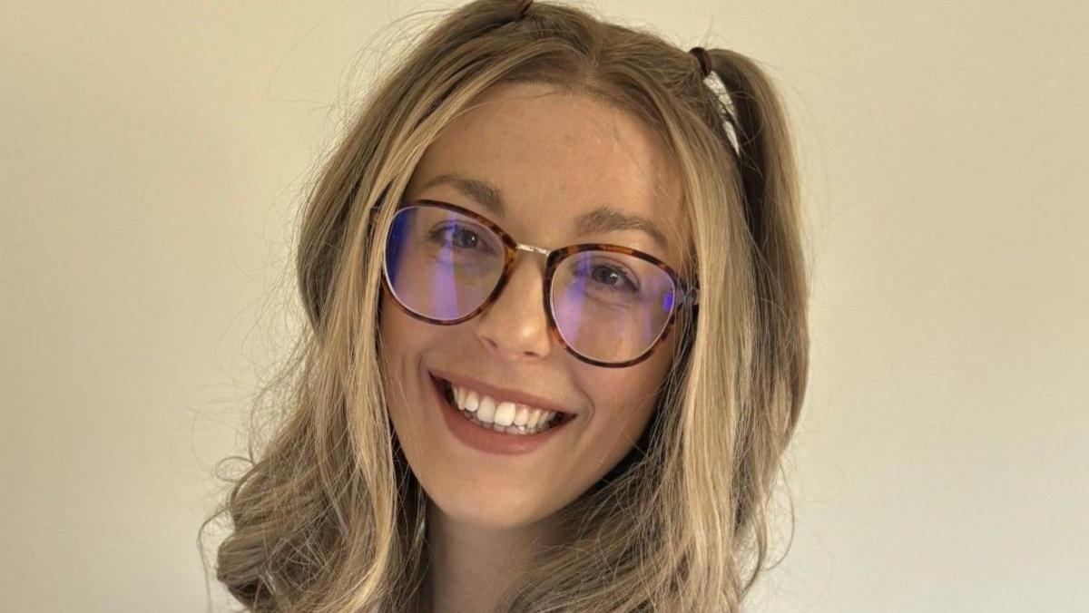 A smiling woman with long blonde hair. She is wearing glasses and posing in front of a cream wall.