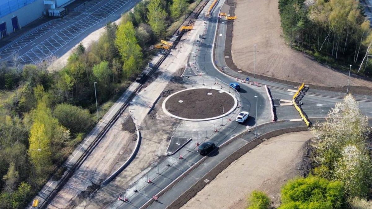 An aerial view of a new roundabout, which has dirt in the middle and white concrete around the circumference. Three partially completed roads link to the roundabout, with work still going on. Trees are visible on either side.