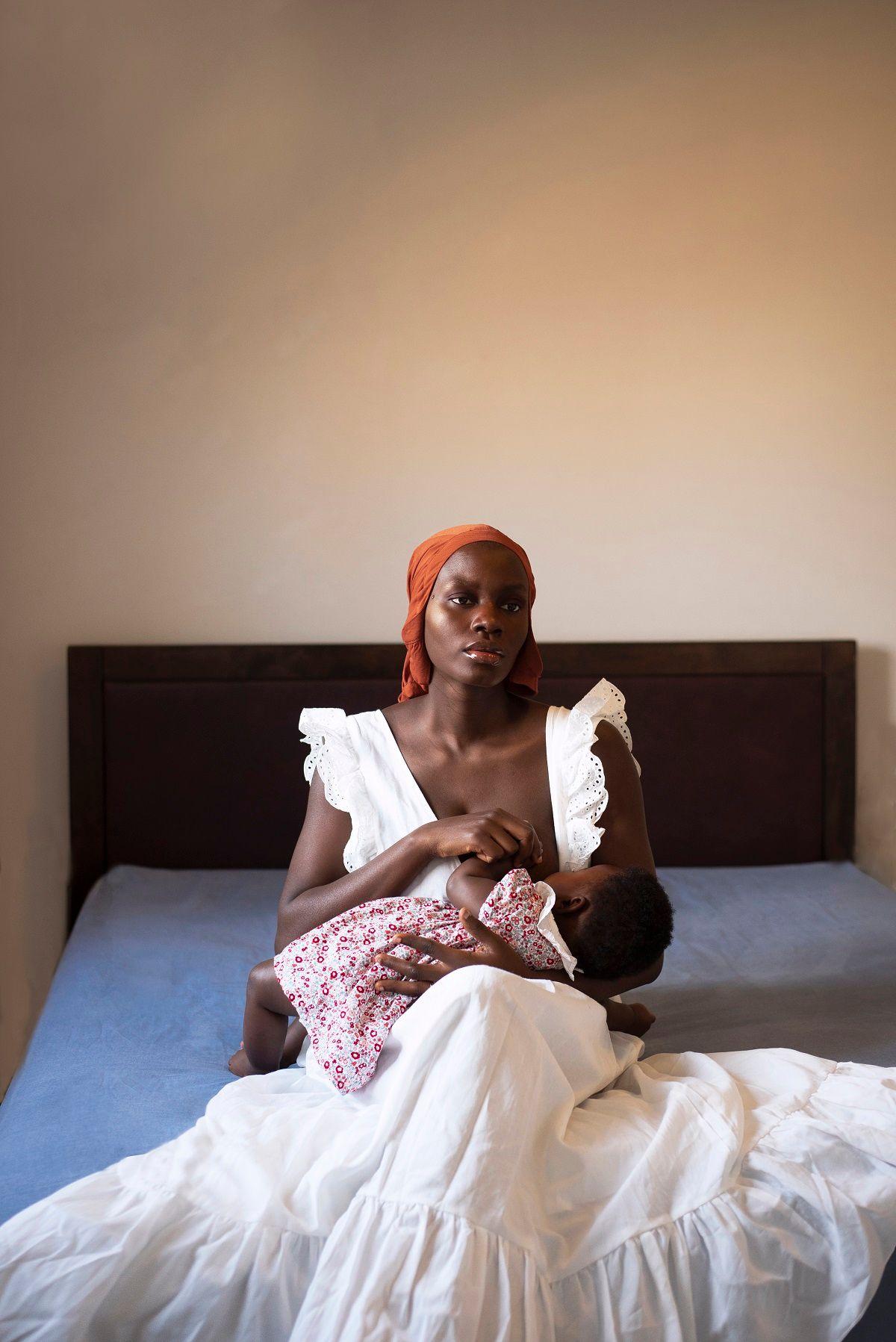 Dola Posh in a red headscarf and white dress is breastfeeding her daughter sitting on the edge of a bed with a blue sheet.