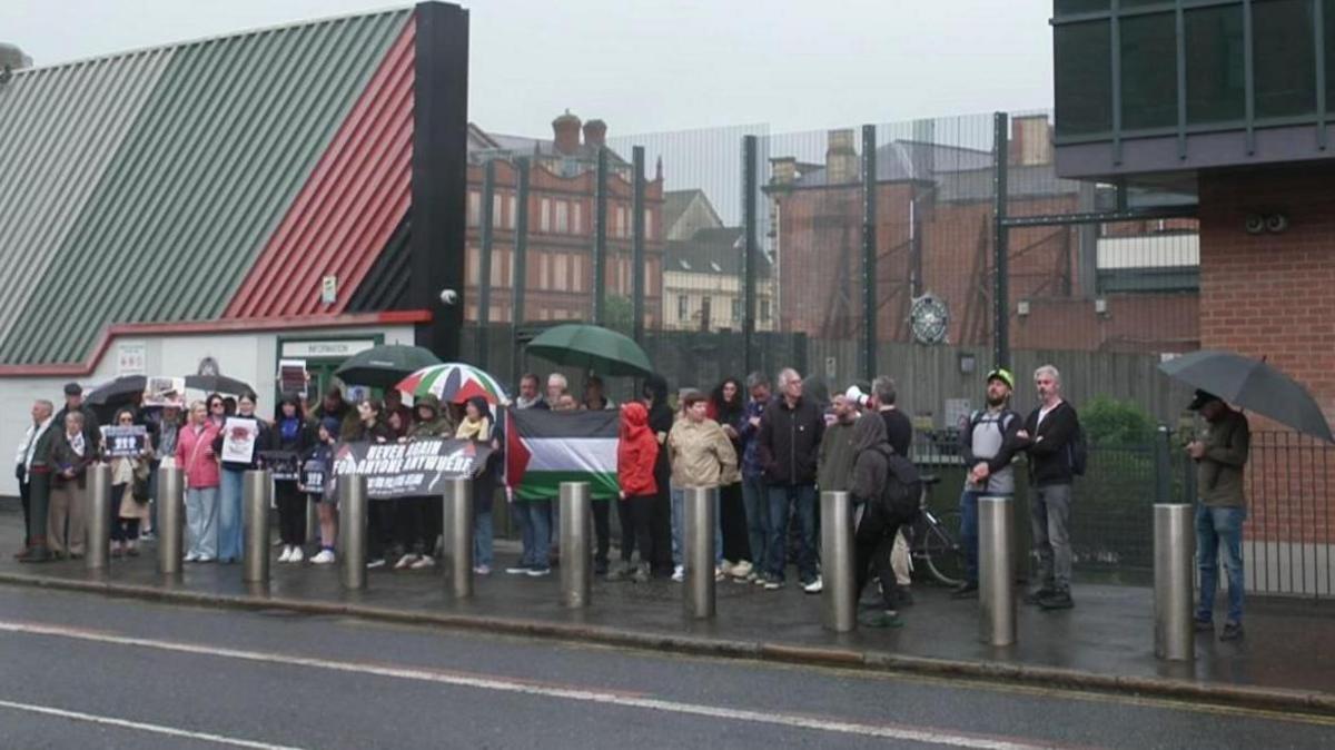 A group of about 30 pro-Palestinian protesters gathered outside Musgrave Street PSNI station in Belfast city centre. Some are holding banners and umbrellas. A large Palestinian flag is being held up in the middle of the group.