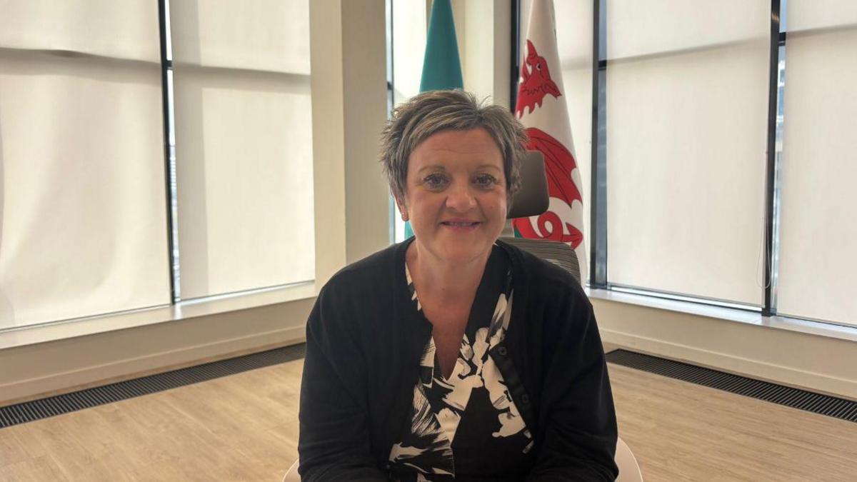 Menyw gyda gwallt golau byr, ffrog ddu a gwyn a siaced ddu yn eistedd tu ol i ddesg gyda baneri Cymru a Kazakhstan tu ôl iddi