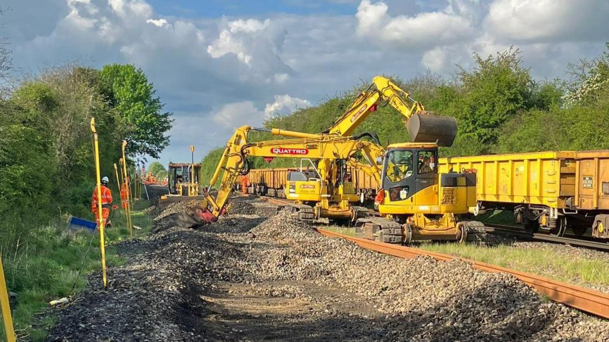 A yellow digger works on a train track as workers in high-visibility clothing and white hard hats stand and watch