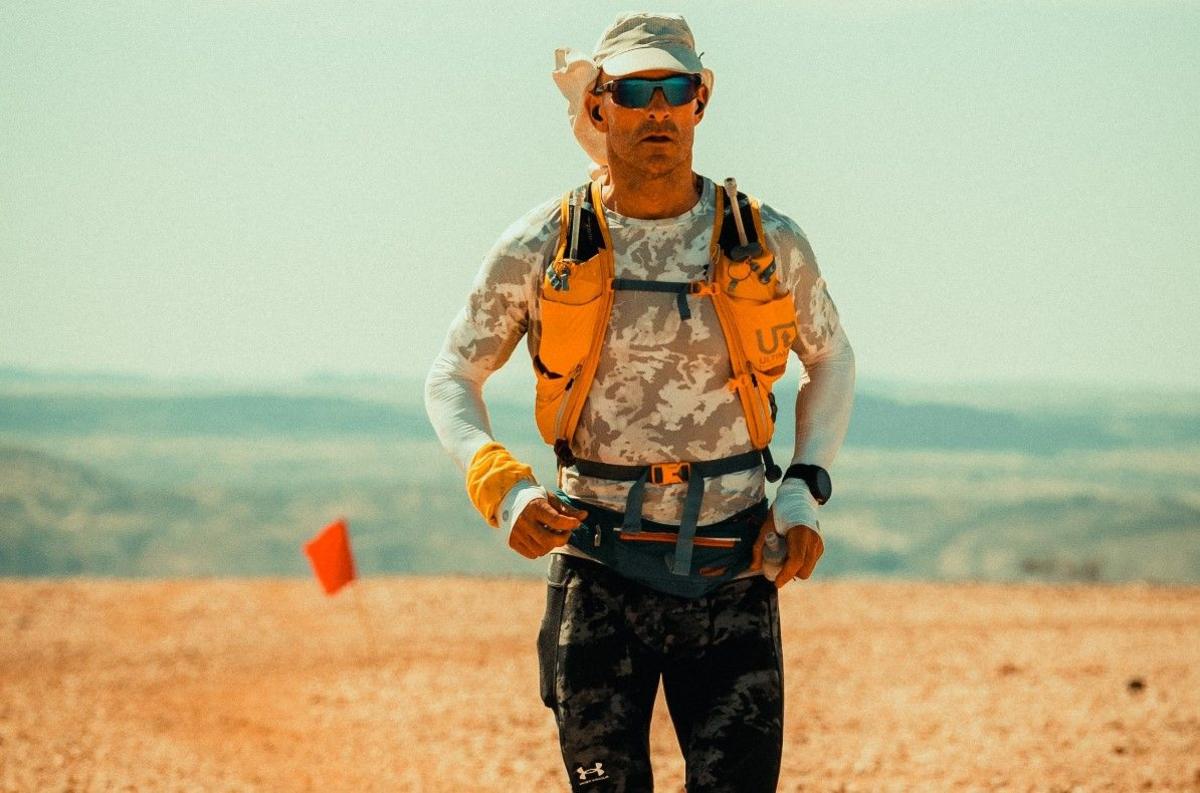 Jon Shield wearing a hat and sunglasses in the desert. A flag is in the ground in the background.