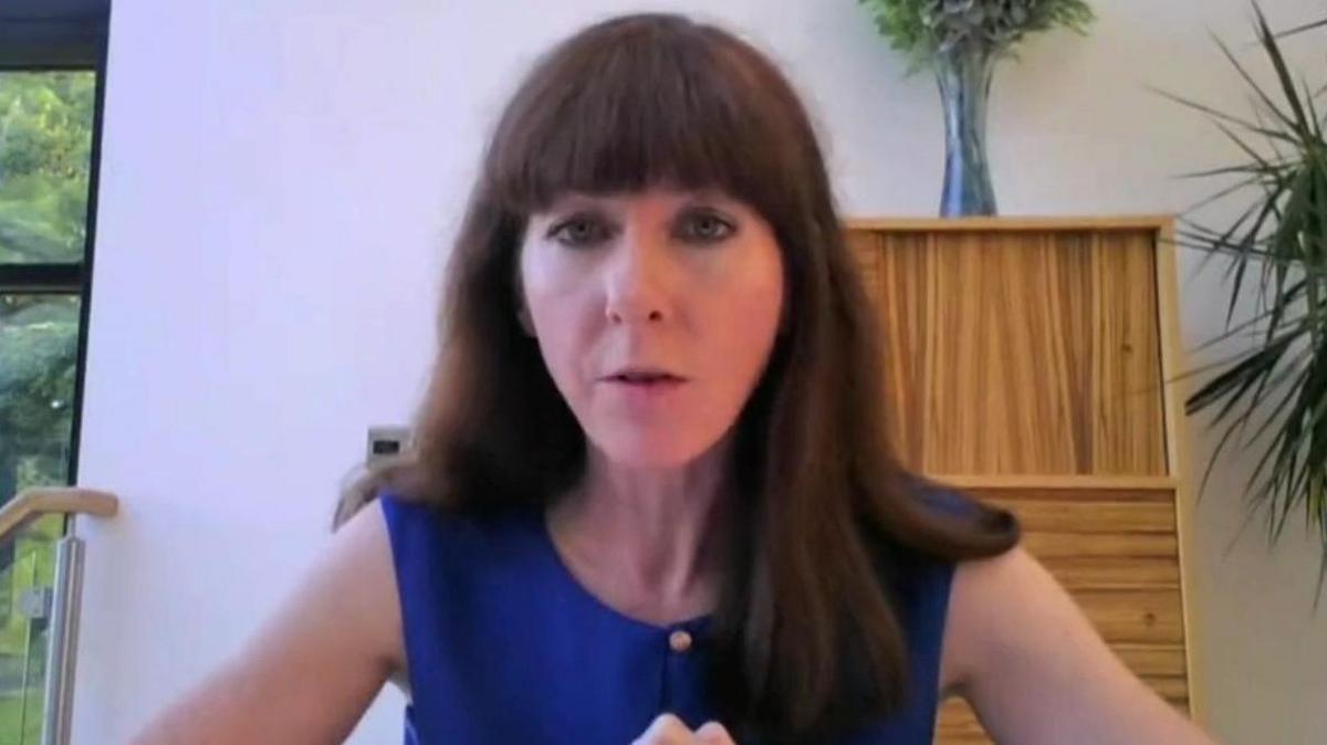 Margaret Farragher has long brown hair with a thick fringe, she is sitting at a desk with light coloured walls and a wooden chest of drawers behind her. She is wearing a short sleeved blue top