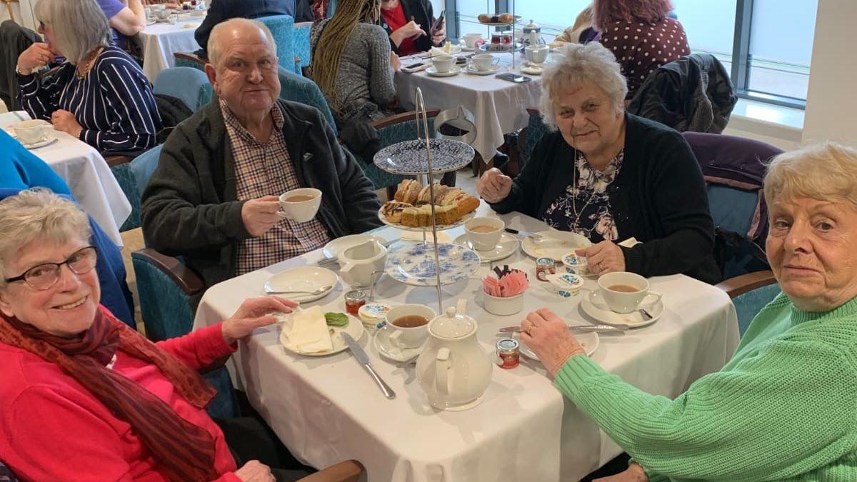 Four elderly people sit around a dinner table drinking tea and eating cake.