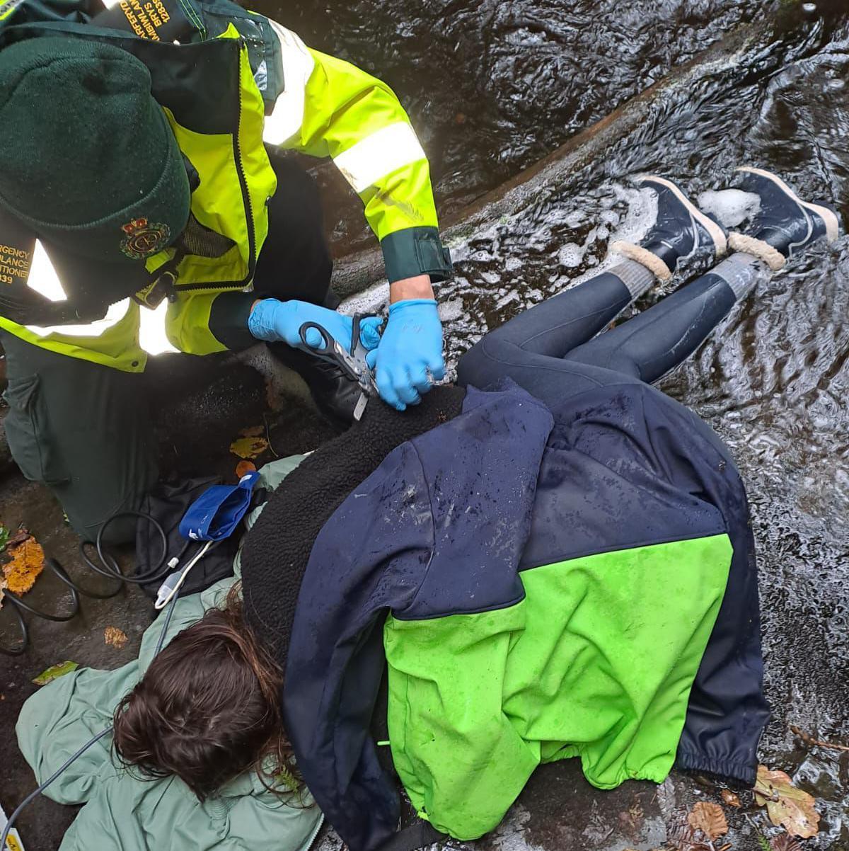 A woman lying on cold wet ground, covered in coats and blankets. Crouched beside her is someone in high viz uniform, wearing a green beanie and blue medical gloves.