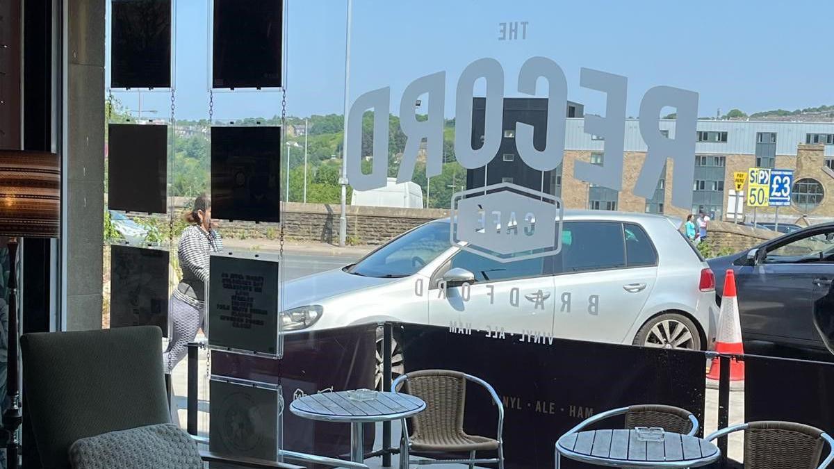 The inside of a bar looking out through a plate glass, floor to ceiling window which shows a silver-coloured car parked close to tables outside. 