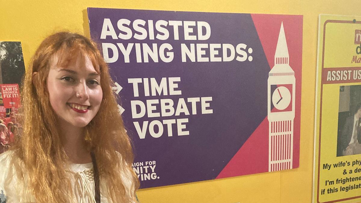 A young woman with red hair standing beside a poster for assisted dying