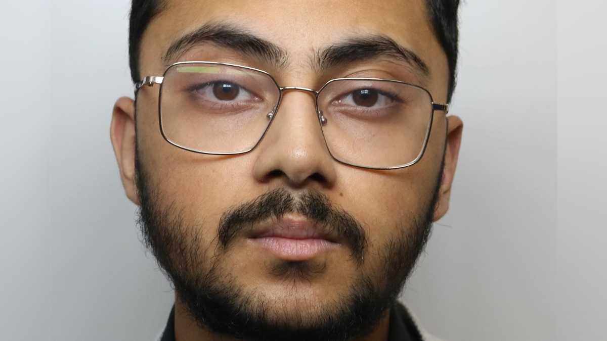 A man with dark hair, dark facial hair and brown eyes. He is wearing glasses. There is a grey background behind him.