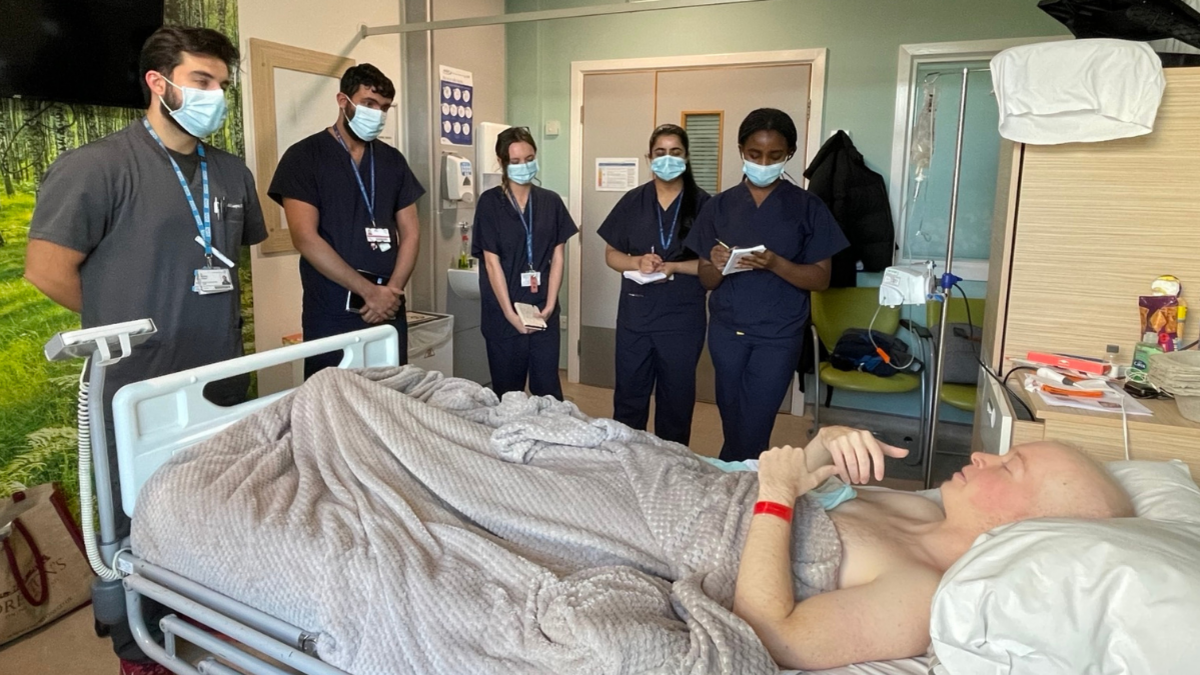 A man in a hospital bed having lost his hair through cancer treatment surrounded by five doctors at the bedside taking notes
