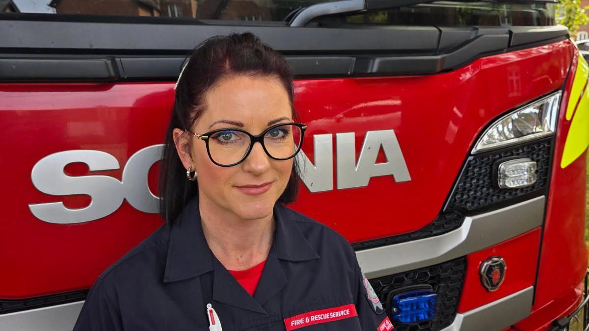A head and shoulder shot of Natalie Clark stood in front of a red fire truck. She is wearing off-duty firefighters uniform and black glasses. She has dark-coloured hair and is smiling at the camera. 
