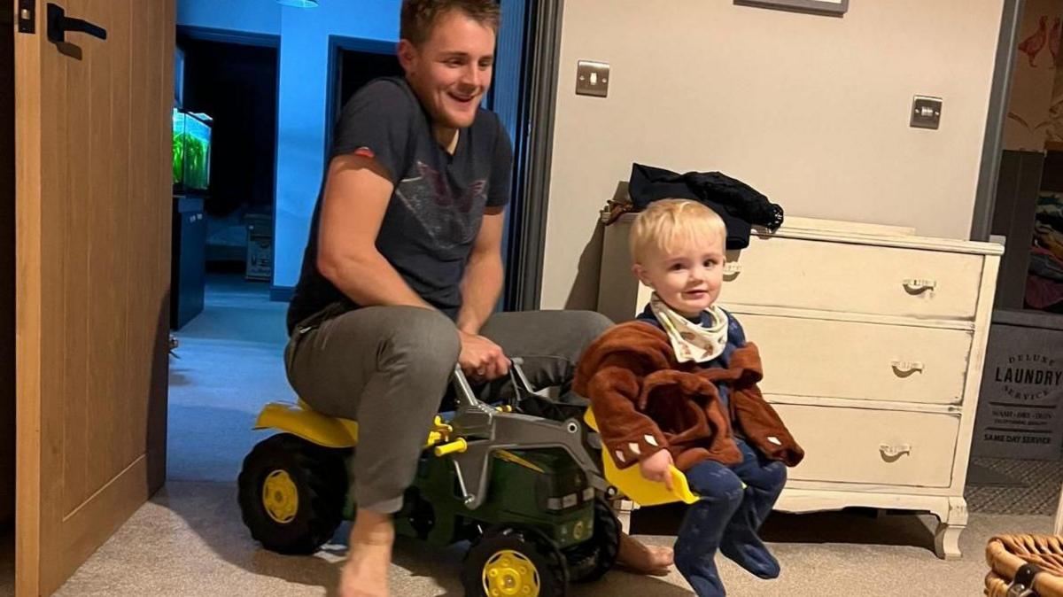 Henry Barringer on a large toy tractor with his son who is sitting in the digger bucket. They are in a bedroom.