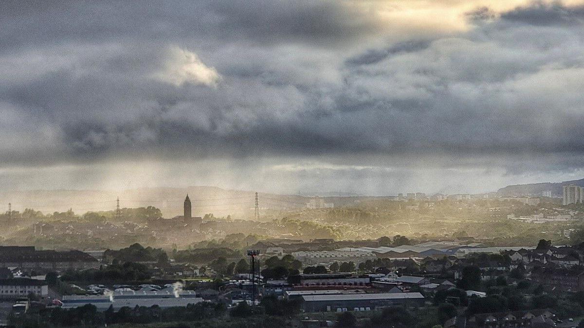Grey clouds with misty rain hang over Glasgow. The city is pictured from far away with many buildings and structures peaking through the mist of the rain.