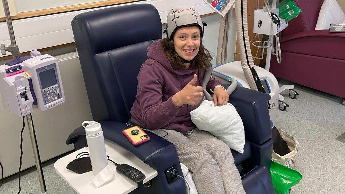 Kat is in a hospital chair. She is wearing a cold cap to help with hair loss during chemotherapy treatment. She is wearing a maroon hoodie and grey trousers. There is a water bottle beside her and hospital equipment.