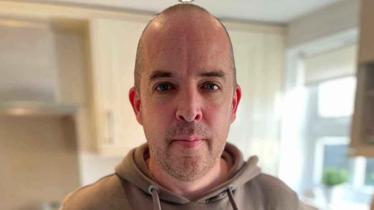 Close up of James Gilmartin who has a shaved head, wearing a light brown hoody, standing in his kitchen