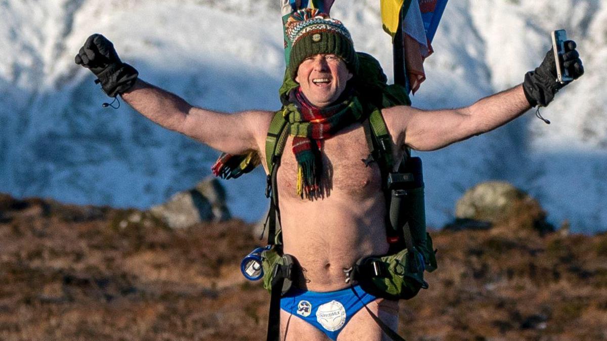 Speedo Mick, dressed in a pair of blue swimwear, gloves, and a rucksack, walking through a mountainous part of the UK with his arms outstretched