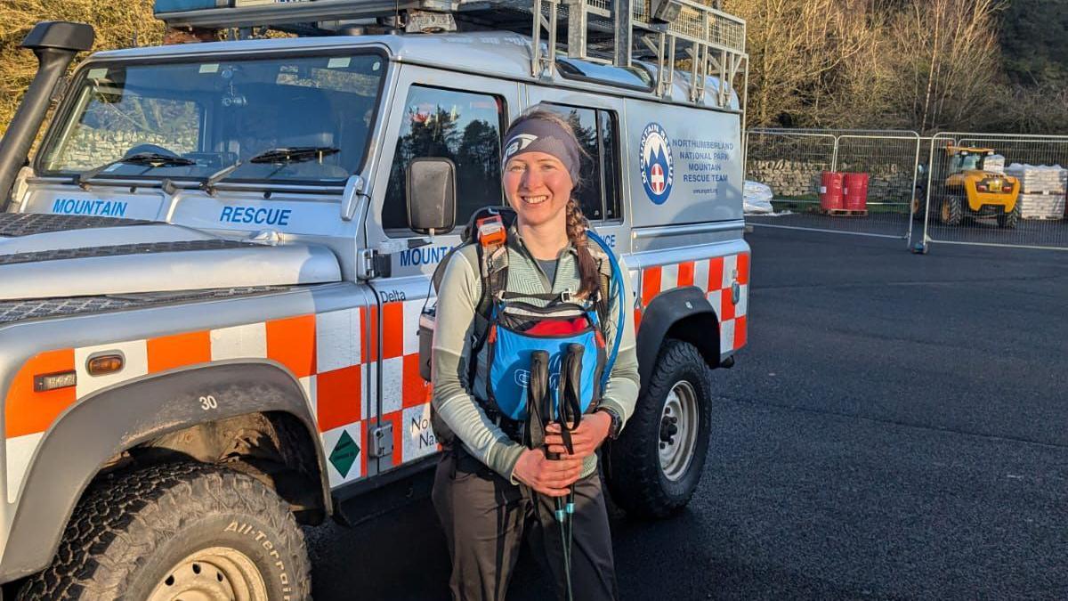 Clëo Bishop-Bolt smiling at the camera in front of a grey and orange Northumberland Mountain Rescue 4x4 car. She's got her long, light brown hair in a plait and is wearing walking-gear, holding two walking poles.