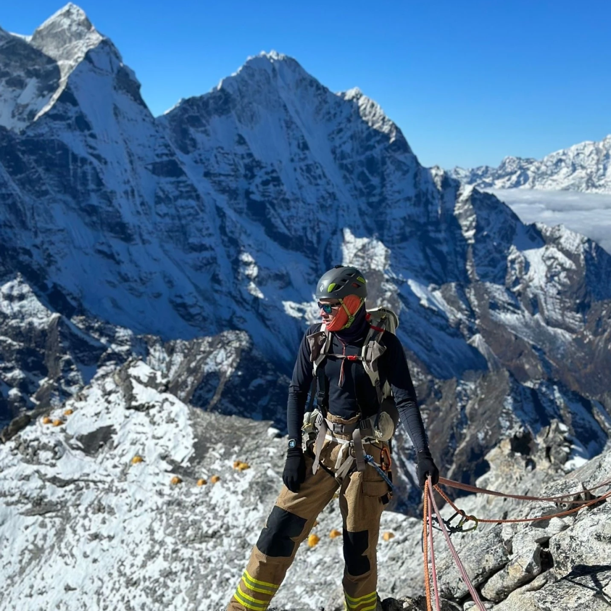 Rhys Fitzgerald standing on a mountain, while holding a rope. In the background are higher mountains, and the sky is a cloudless blue