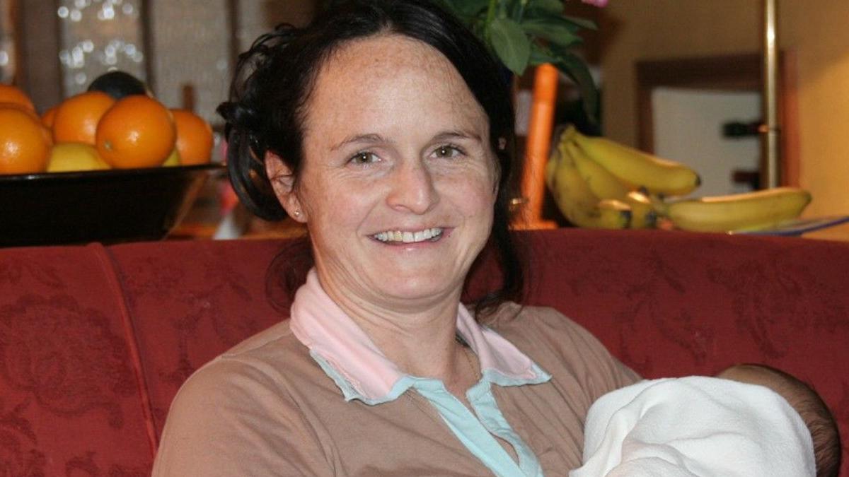 Sammy Young, pictured smiling and holding a baby. She has dark brown hair which is tied up, and she has freckles on her forehead. She is wearing a brown top with a pink collar and sitting on a red patterned sofa. In the background, on a table, is a bowl of oranges and some bananas.