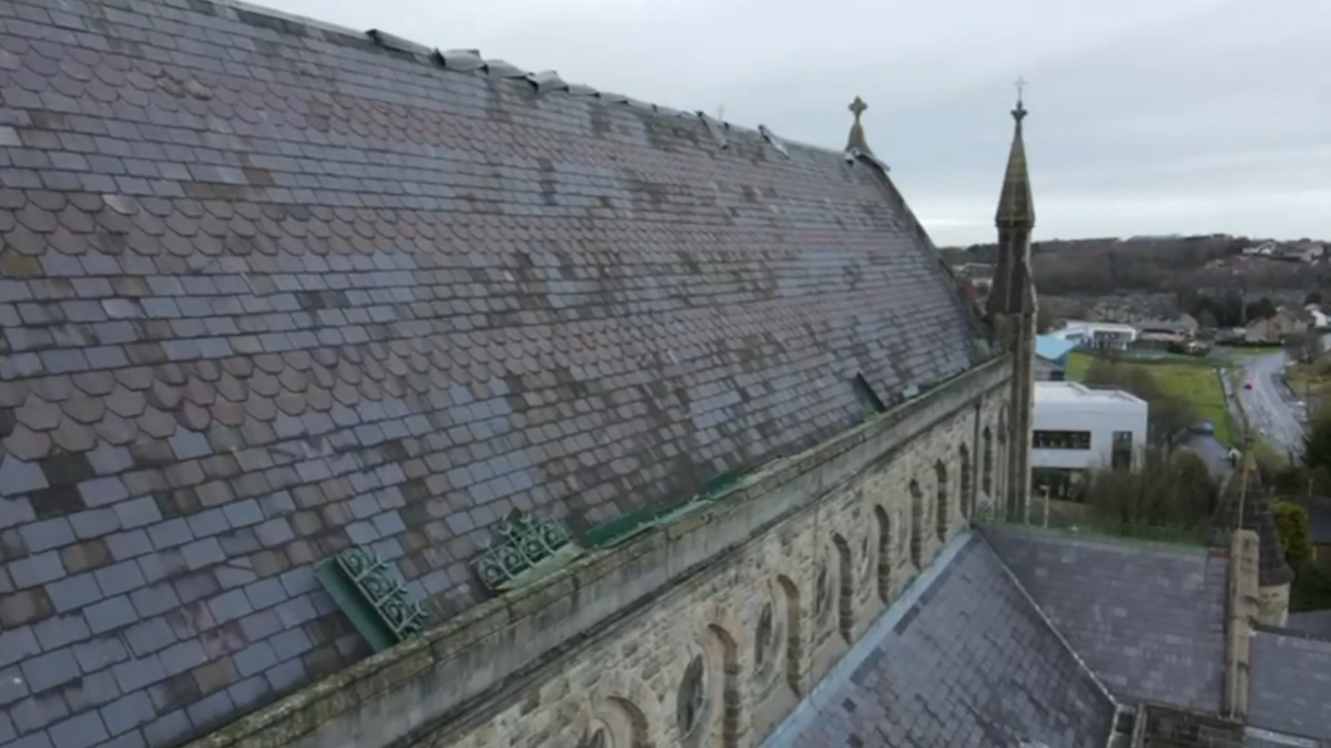 Aerial image of the slanted main roof of a church. We can see grey roof slates, some of which have clearly been displaced. The shot shows a full side profile of the church roof, with the front of the church towards the back of the imae. We an also see a landscape beyond the church - some building, a road and a hill.