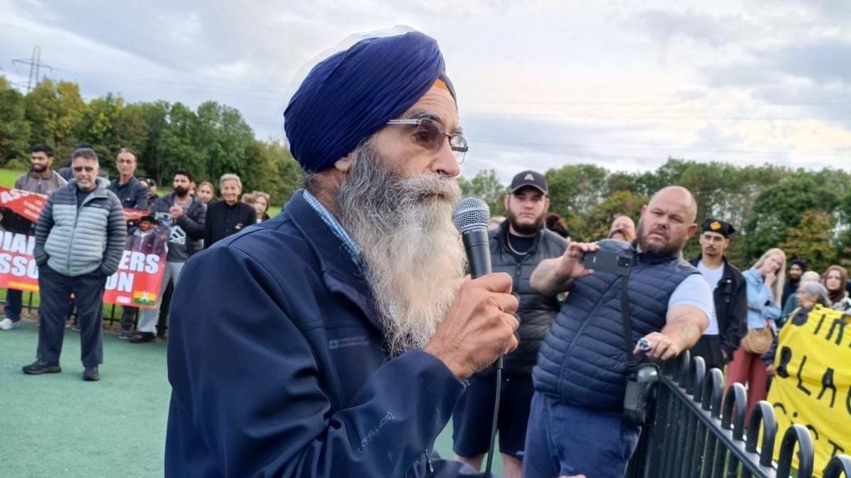 A man wearing a blue turban, blue jacket and with a long grey beard holds a microphone. He is flanked by people taking pictures and security