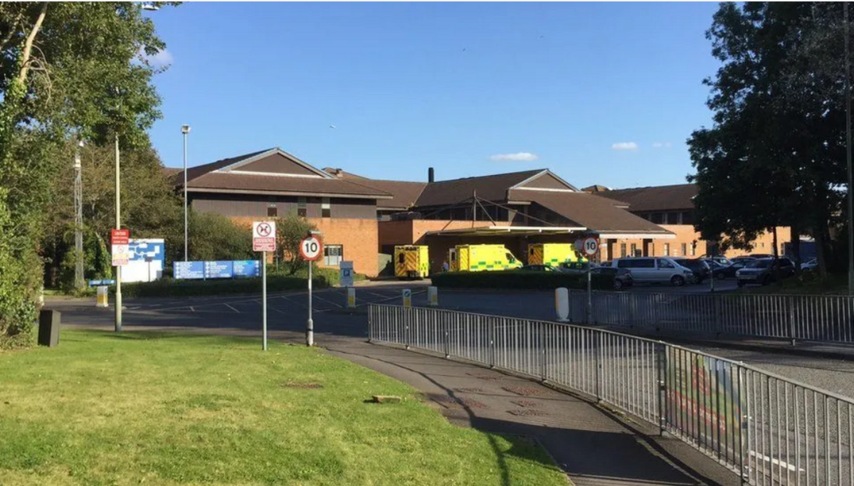 The brown exterior of the Princess of Wales hospital building, with three ambulances and several cars parked outside, welcome and road signs to the left and a patch of grass in front.