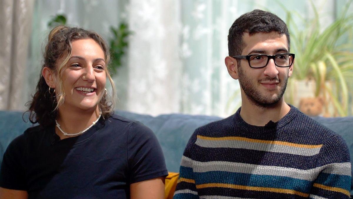 Aviva and Eric sitting on a blue sofa with white net curtains and a spider plant in the background. Aviva, on the left, is smiling and wear a black short-sleeved top with a white beaded necklace. She has gold hoops and her shoulder-length hair is half tied-up, with two strands of hair framing her face. Eric, on the left, is smiling slightly and wears a striped jumper and dark rimmed glasses. He has very dark, short hair. The siblings are both looking off to the right, beyond the camera. 