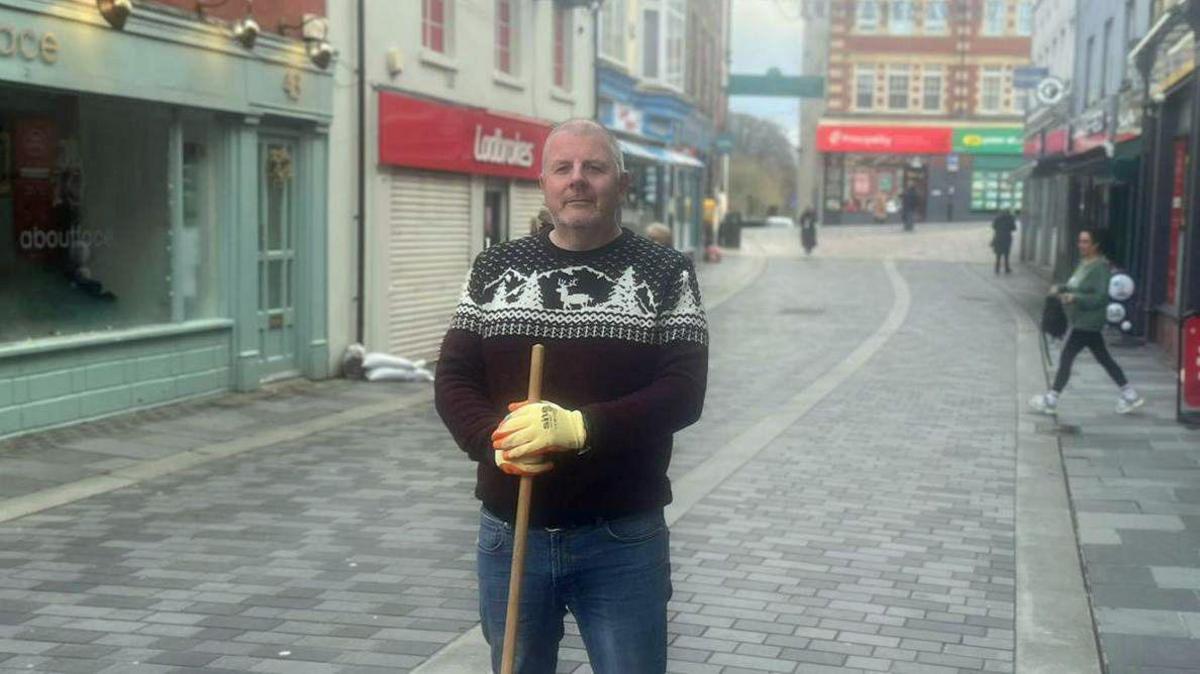 Jeff Baxter holding a sweeping brush and wearing gloves as he stands on Mill Street 