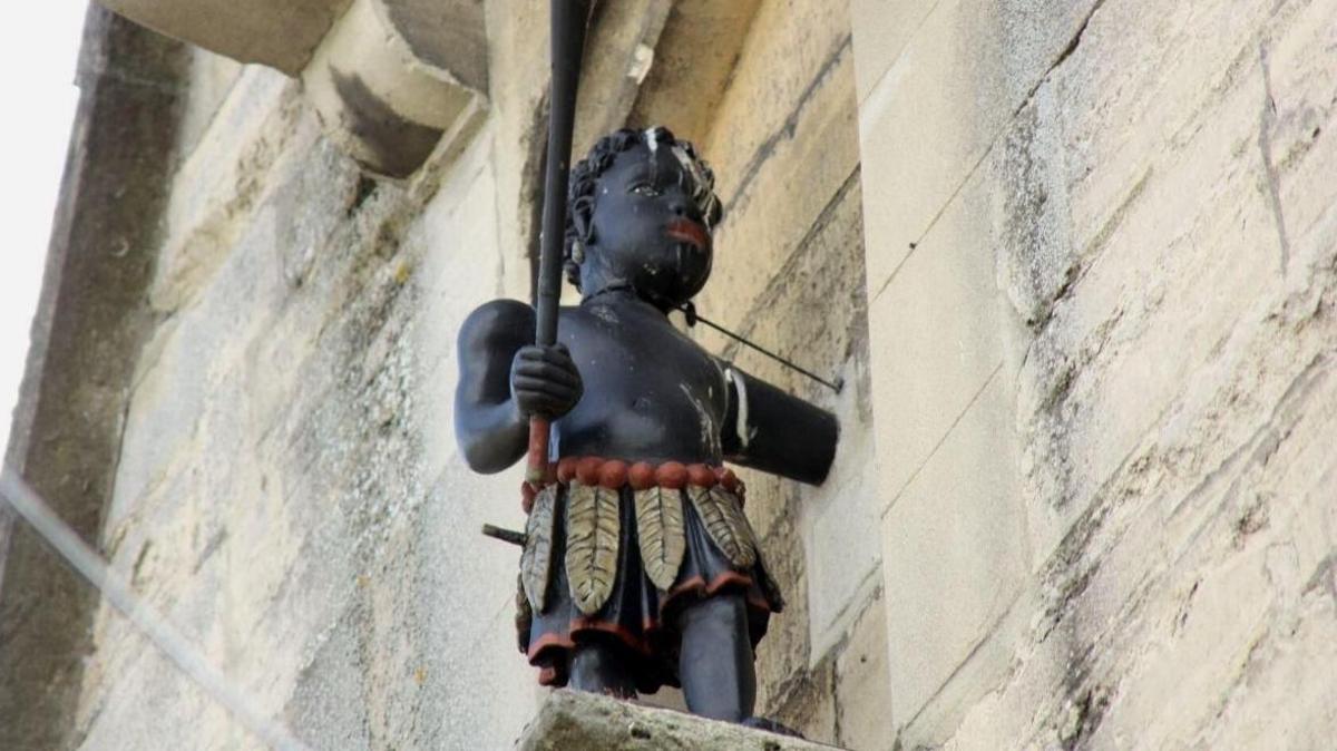 The Blackboy statue on an old church building in Stroud. It is a black statue depicting a young boy wearing a skirt made of feathers and carrying a pole to ring the bell with. He has a wire around his neck attaching him to the wall.